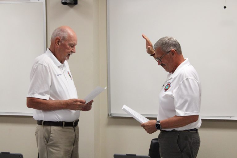 Two men in white shirts stand facing each other in a classroom, holding papers. One man raises his right hand as if taking an oath, while the other looks on and reads from his paper. Whiteboards are visible in the background.