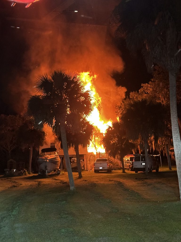 A large fire engulfs a building at night, with flames rising behind palm trees and parked vehicles in the foreground.