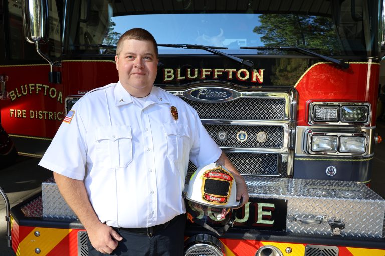 A Bluffton fire battalion chief stands in front of a red fire engine holding his white fire helmet.