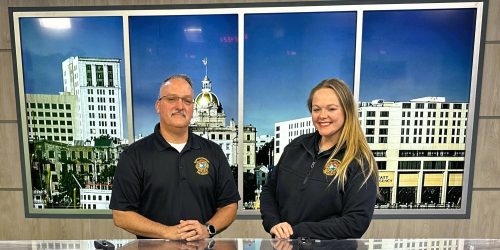 Two people in black polo shirts with official logos stand behind a news desk, smiling, in front of a backdrop showing city buildings and a gold-domed structure.